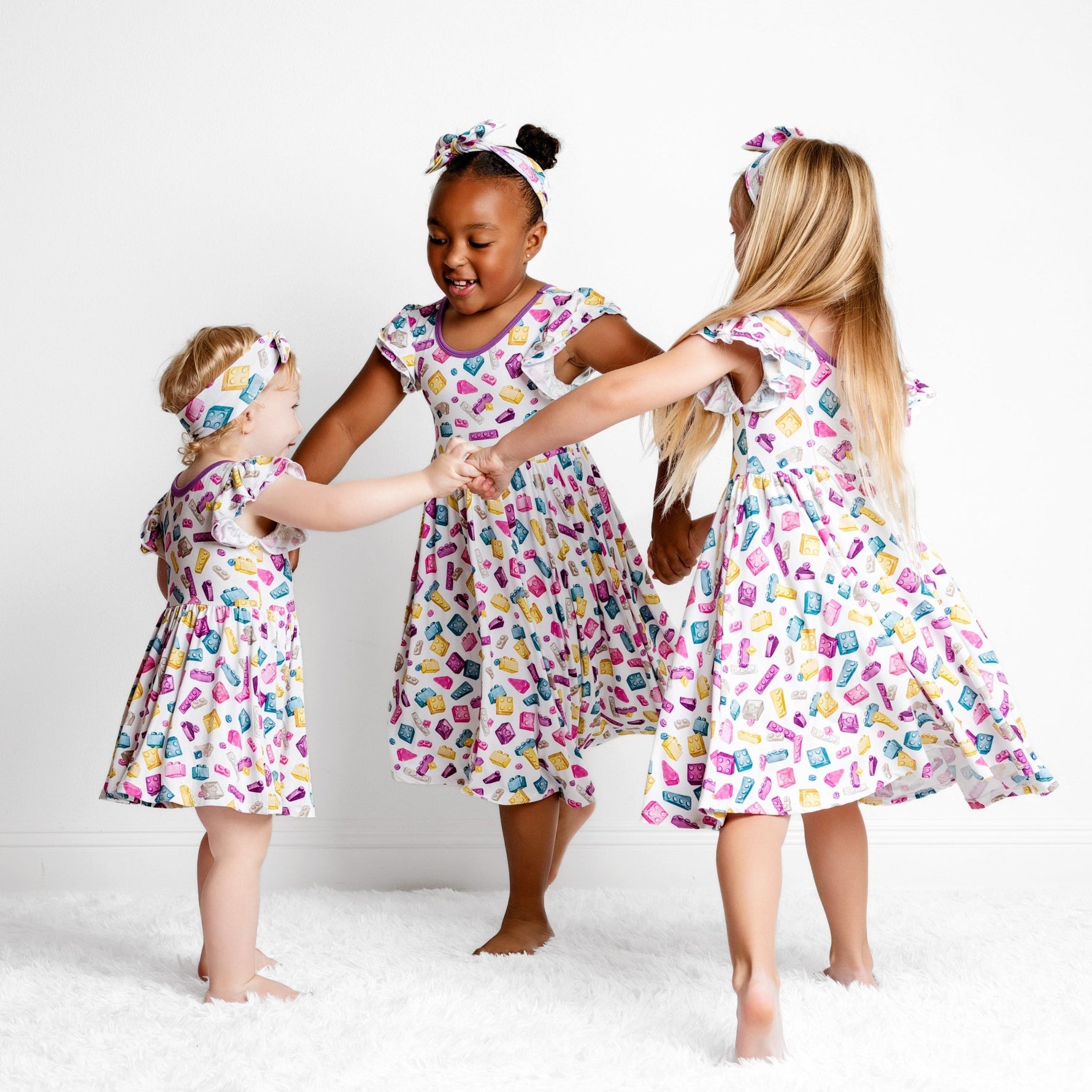 Three young girls wearing Big Dreams Little Jammies' Block Party (Purple) Short Sleeve Big Kid Twirl Dress hold hands and dance in a circle on a white, fluffy rug against a plain white background.