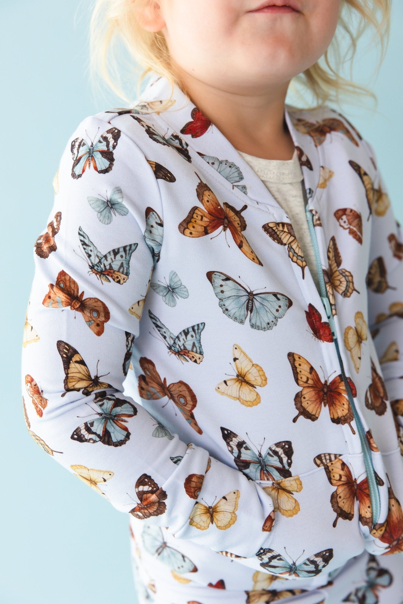 A child models the Jammie Session Butterfly Kisses Kids' Jacket—featuring colorful butterfly prints—against a light blue background.