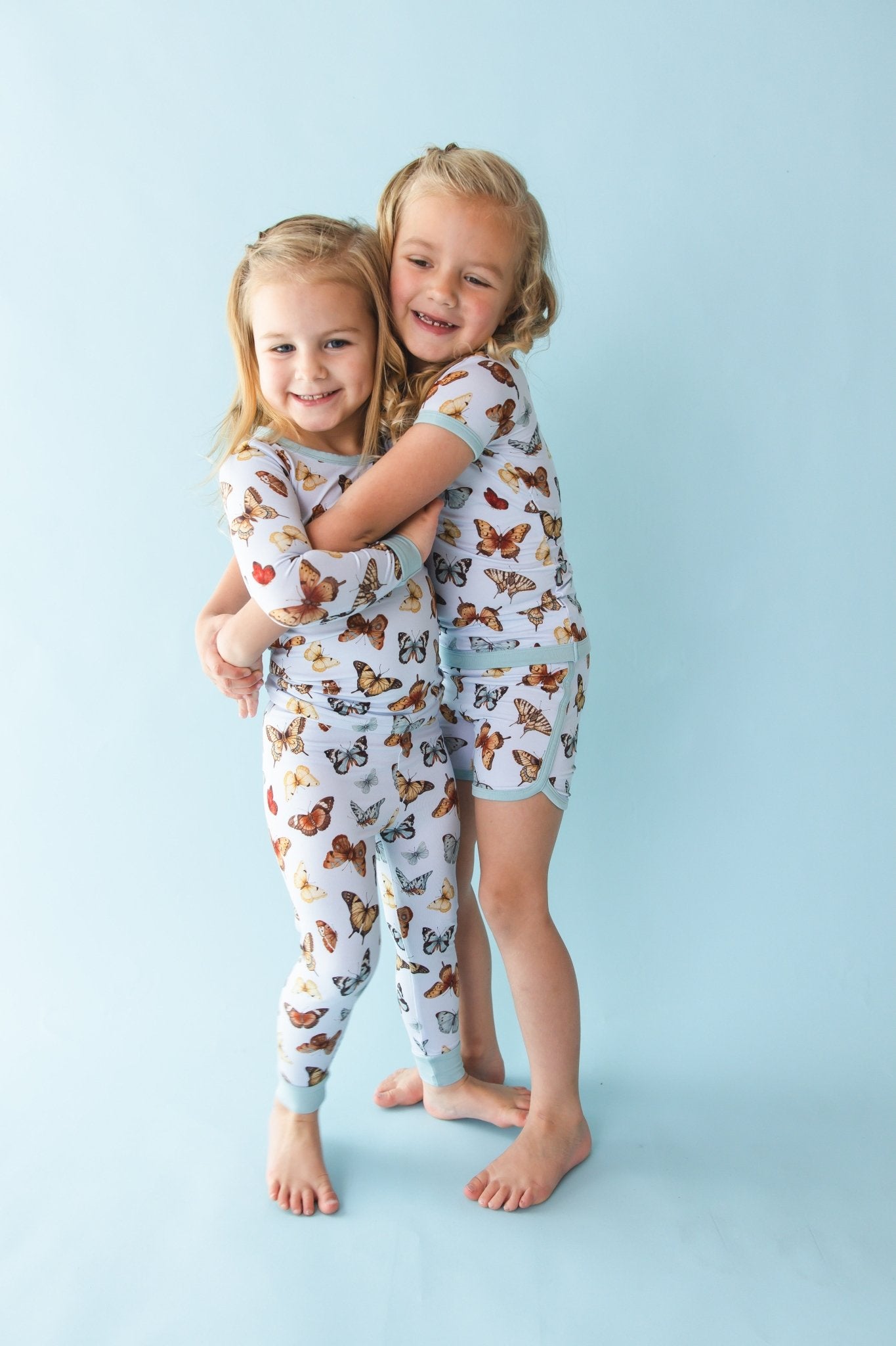 Two young girls smile and hug on a light blue background, both wearing matching Jammie Session Butterfly Kisses Short Pajama Sets featuring a playful butterfly pattern.