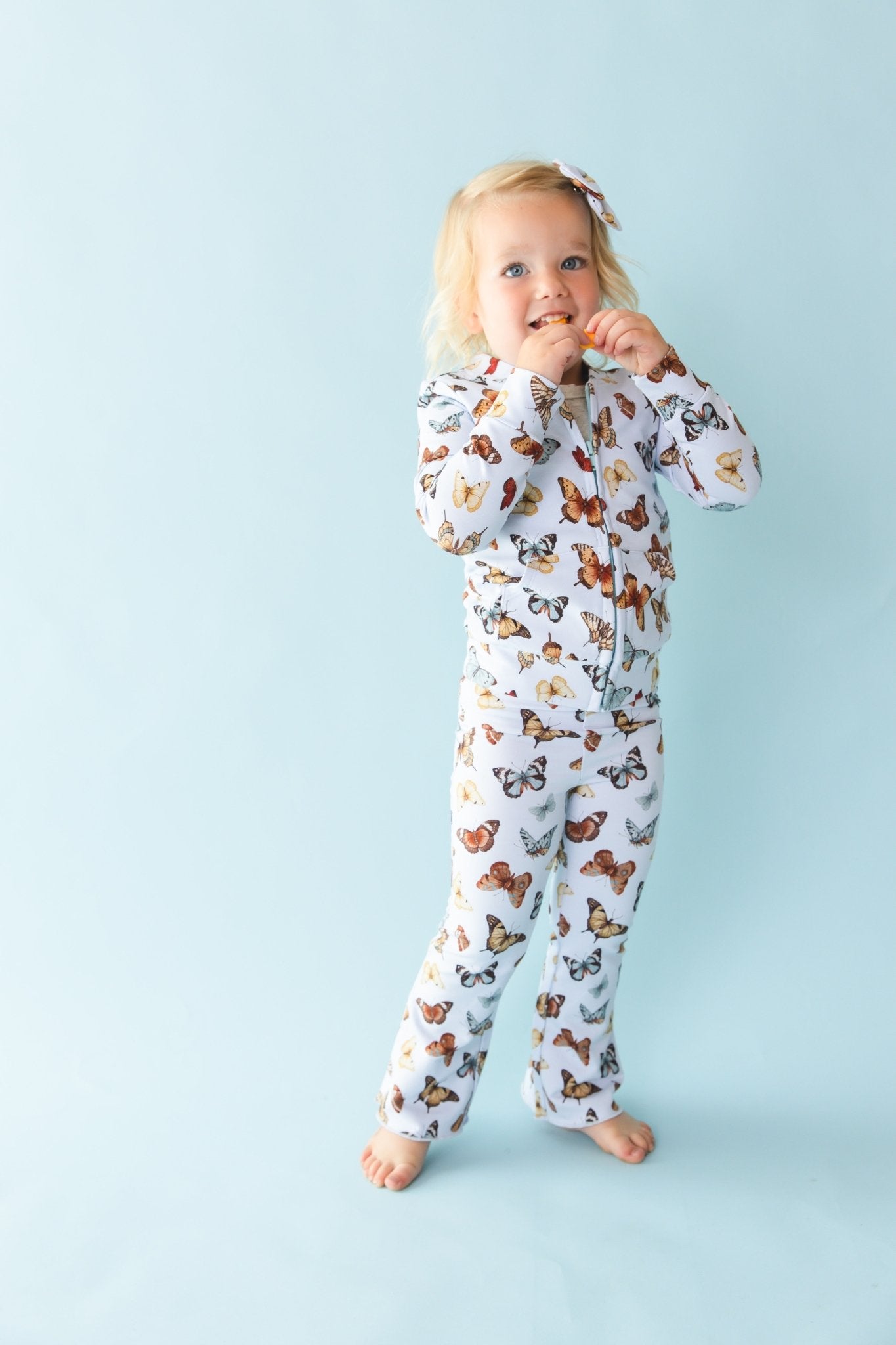 A young child with blonde hair stands barefoot against a light blue background, wearing Jammie Session's Butterfly Kisses Kids' Flare Pants and a matching hair bow, made from breathable bamboo cotton blend fabric.
