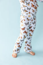 A child is wearing Jammie Session’s Butterfly Kisses Kids' Flare Pants—breathable white pajamas with a colorful butterfly print, shown barefoot against a light blue background, lower body only visible.