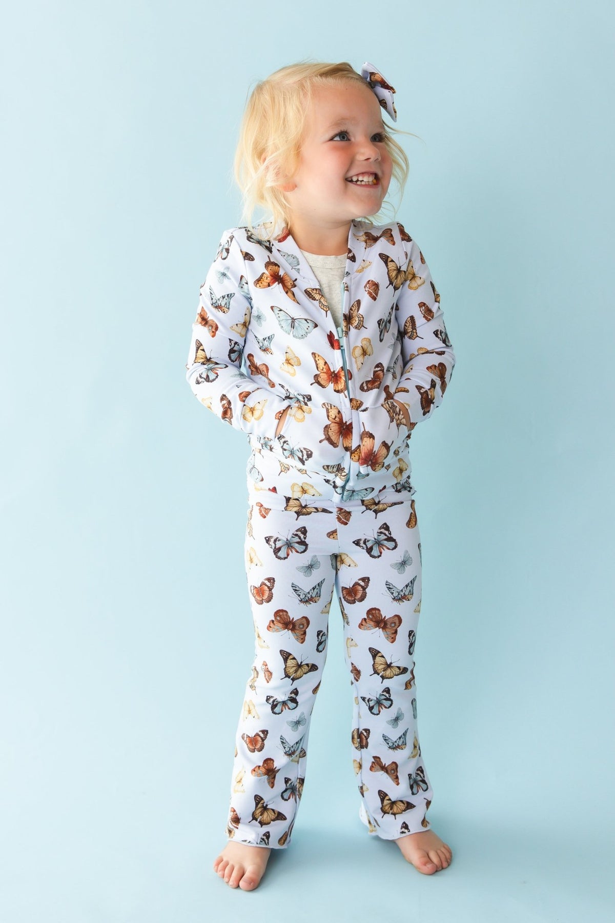 A young child smiles barefoot against a light blue background, wearing Jammie Session’s Butterfly Kisses Kids' Flare Pants—hypoallergenic and crafted from a soft bamboo cotton blend.