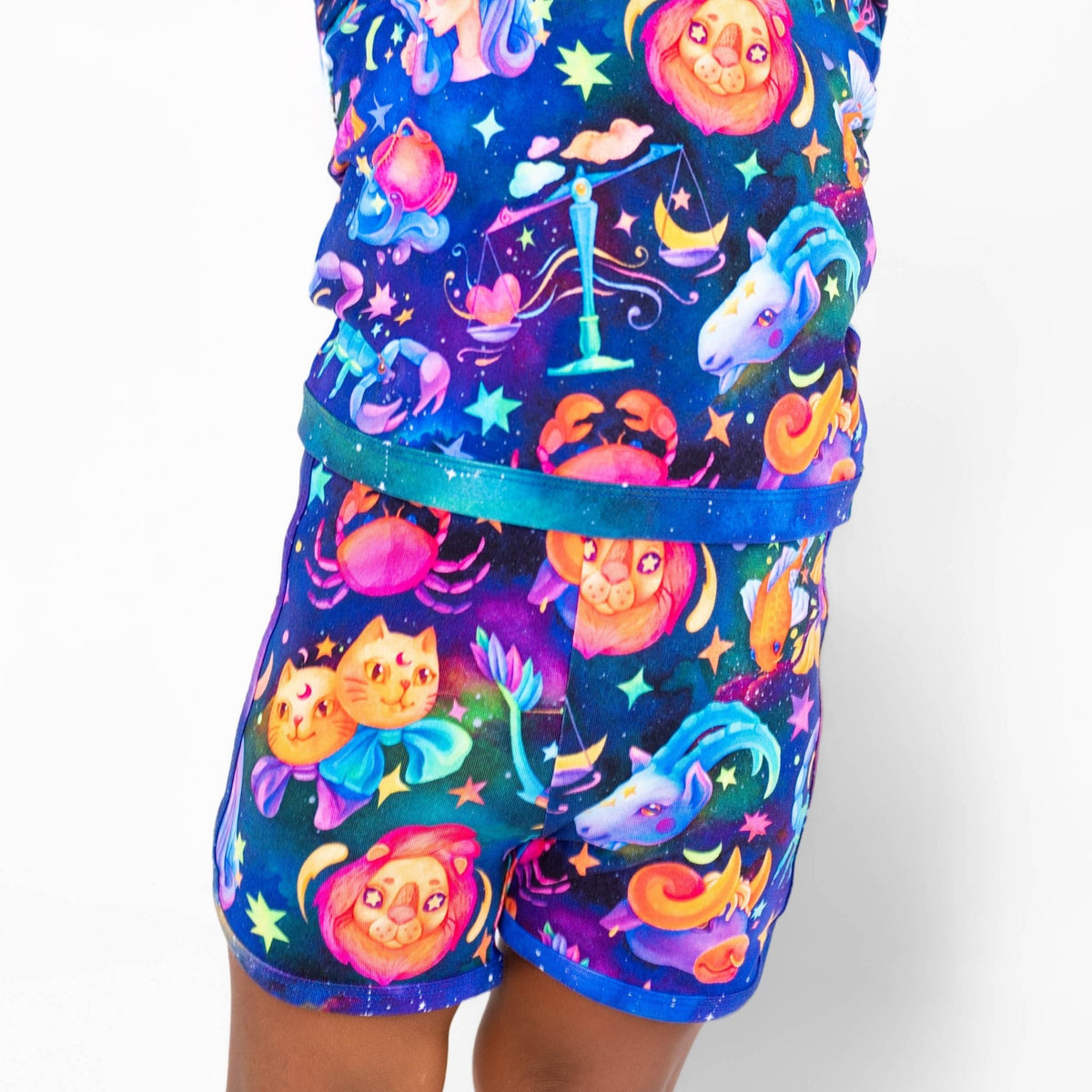 A child wears the Jammie Session Cosmic Cuddles Zodiac Short Pajama Set, crafted from buttery soft, hypoallergenic bamboo and adorned with lions, crabs, unicorns, and stars on a dark blue background.