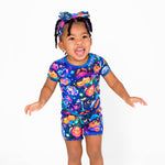 A young child wears the buttery soft, hypoallergenic Cosmic Cuddles Zodiac Short Pajama Set by Jammie Session in blue, paired with a colorful Halloween-themed headband, standing against a plain white background.