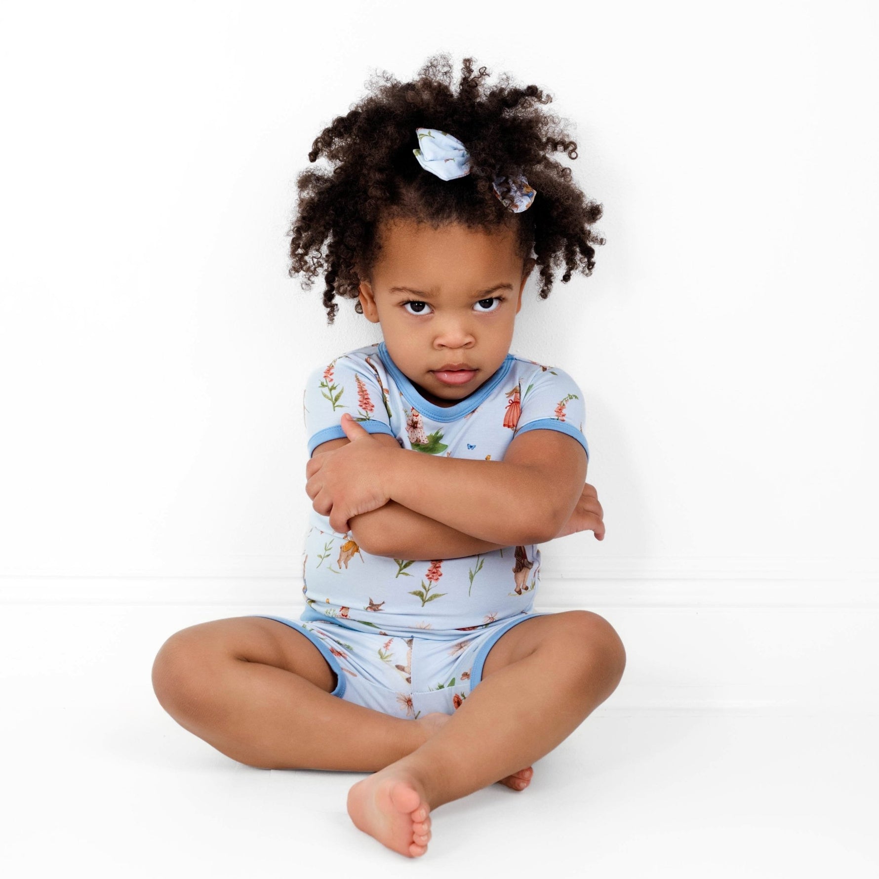 A young child with curly hair sits on the floor, arms crossed, wearing the Jammie Session "Don’t Worry, Be Hoppy (Rabbit)" Short Pajama Set and a matching bow, looking seriously at the camera.