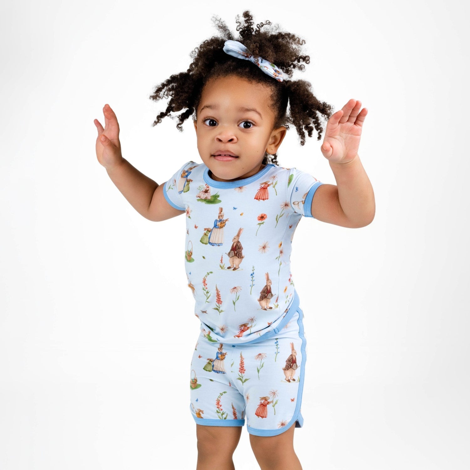 A young child in the Jammie Session “Don’t Worry, Be Hoppy (Rabbit)” blue bamboo short pajama set with playful illustrations stands with raised arms against a plain white background.