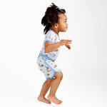 A young child with curly hair stands barefoot against a plain white background, wearing the Jammie Session “Don’t Worry, Be Hoppy (Rabbit)” Short Pajama Set featuring light blue and colorful prints.