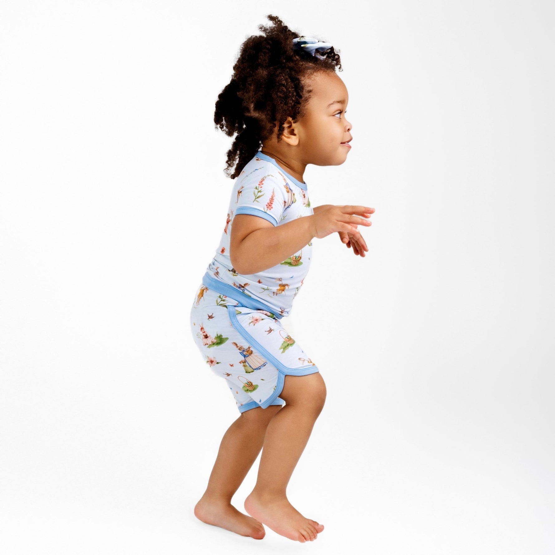 A young child with curly hair stands barefoot against a plain white background, wearing the Jammie Session “Don’t Worry, Be Hoppy (Rabbit)” Short Pajama Set featuring light blue and colorful prints.
