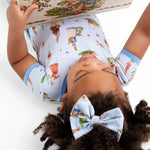 A child with curly hair and a matching bow reads while lying down, cozy in the Jammie Session “Don’t Worry, Be Hoppy (Rabbit)” short pajama set—soft bamboo fabric that’s gentle on sensitive skin, hypoallergenic, and moisture-wicking.