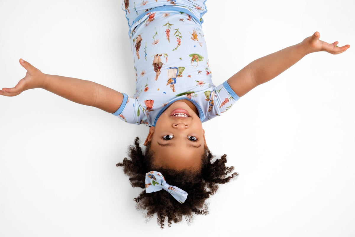 A young child smiles and lies on their back with arms outstretched, wearing the Jammie Session “Don’t Worry, Be Hoppy (Rabbit)” short pajama set and matching headband, photographed from above on a white background.