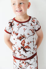 A young child wears the Jammie Session Dreaming with Dragons Short Pajama Set—breathable kids’ pajamas featuring a matching short-sleeve top and shorts with a brown dragon and floral print—posed against a white background.