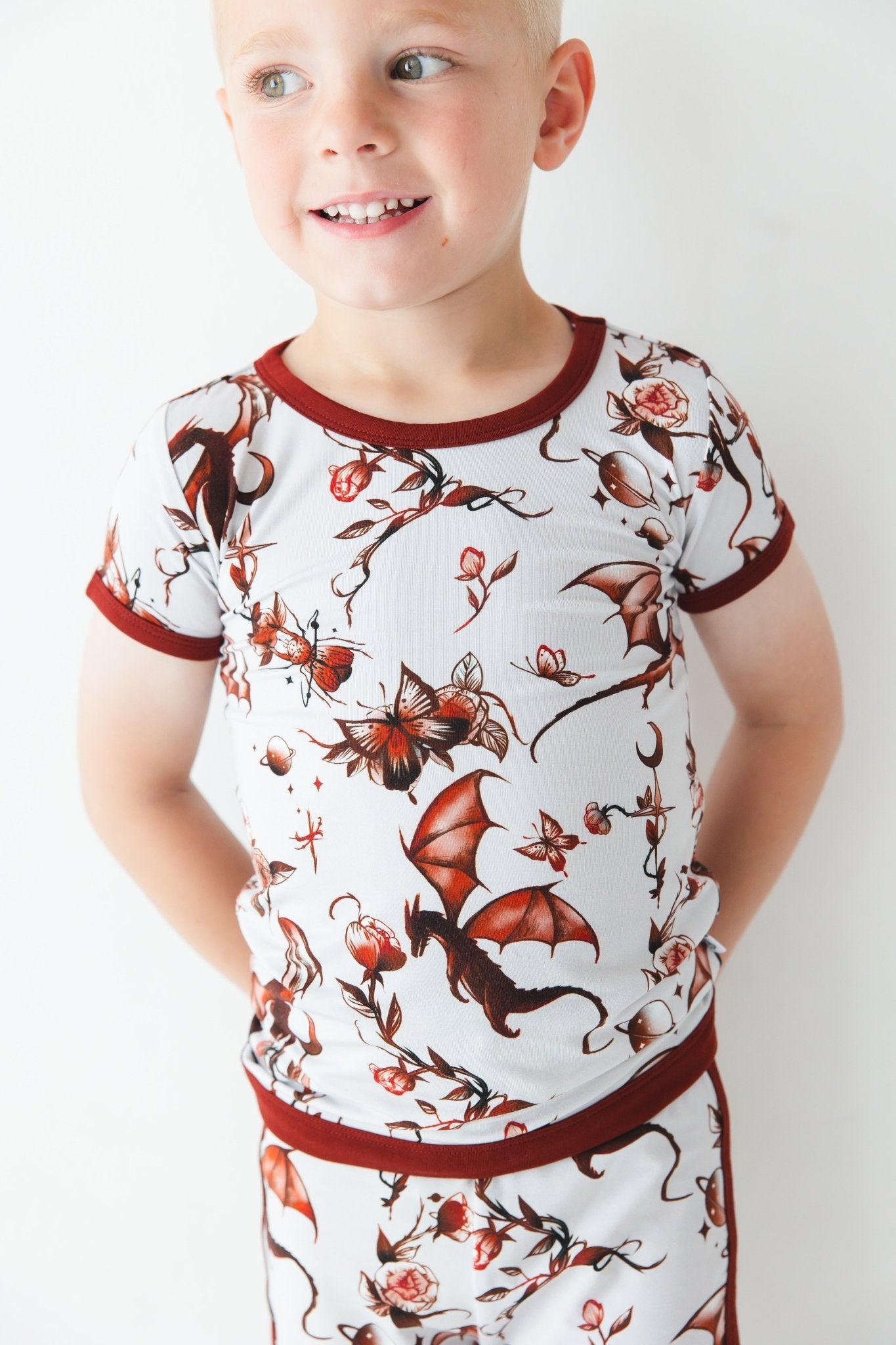 A young child wears the Jammie Session Dreaming with Dragons Short Pajama Set—breathable kids’ pajamas featuring a matching short-sleeve top and shorts with a brown dragon and floral print—posed against a white background.