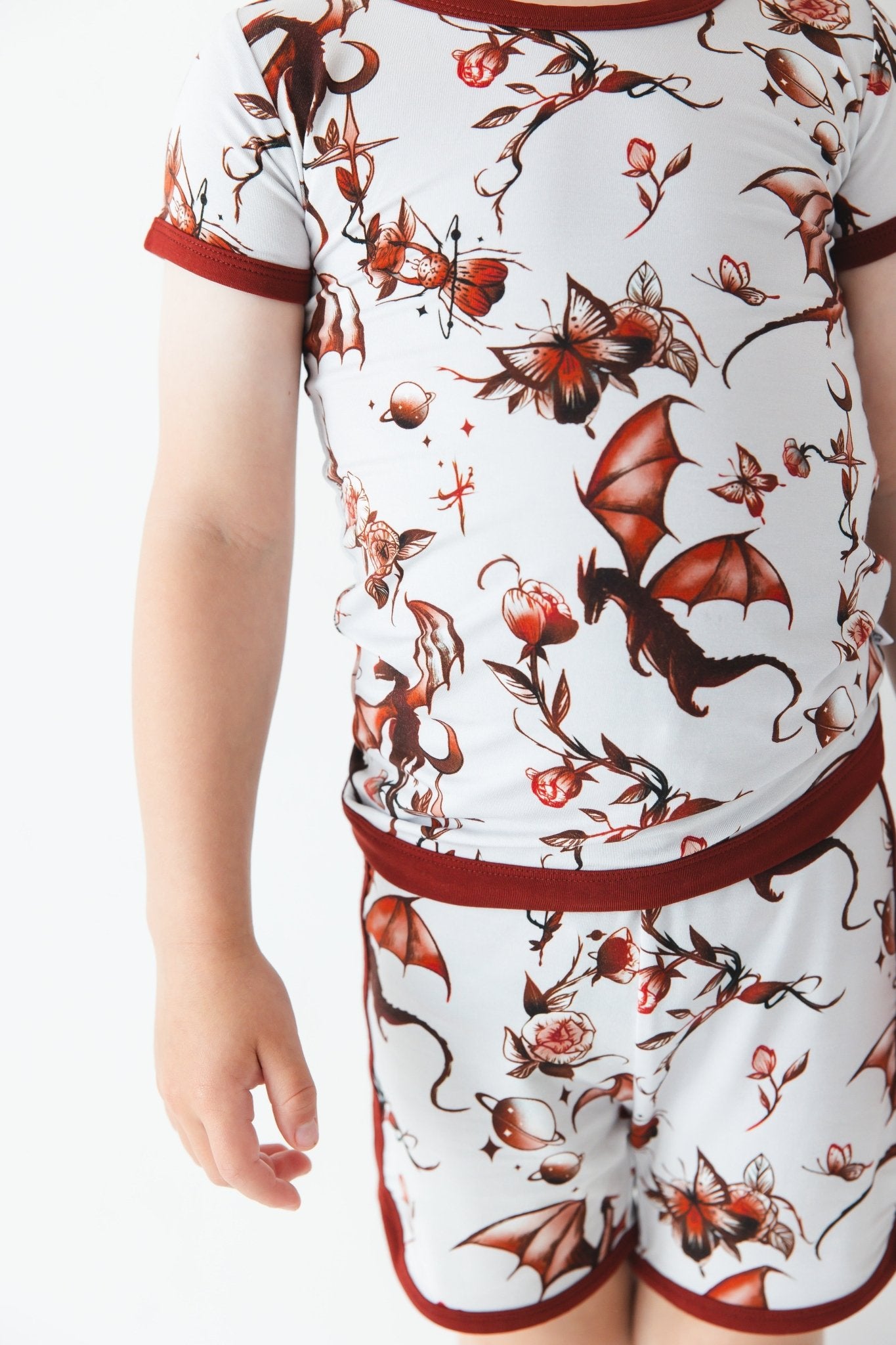 A child in the Jammie Session Dreaming with Dragons Short Pajama Set—featuring a matching short-sleeve shirt and shorts with red dragon and floral prints—stands against a plain white background.