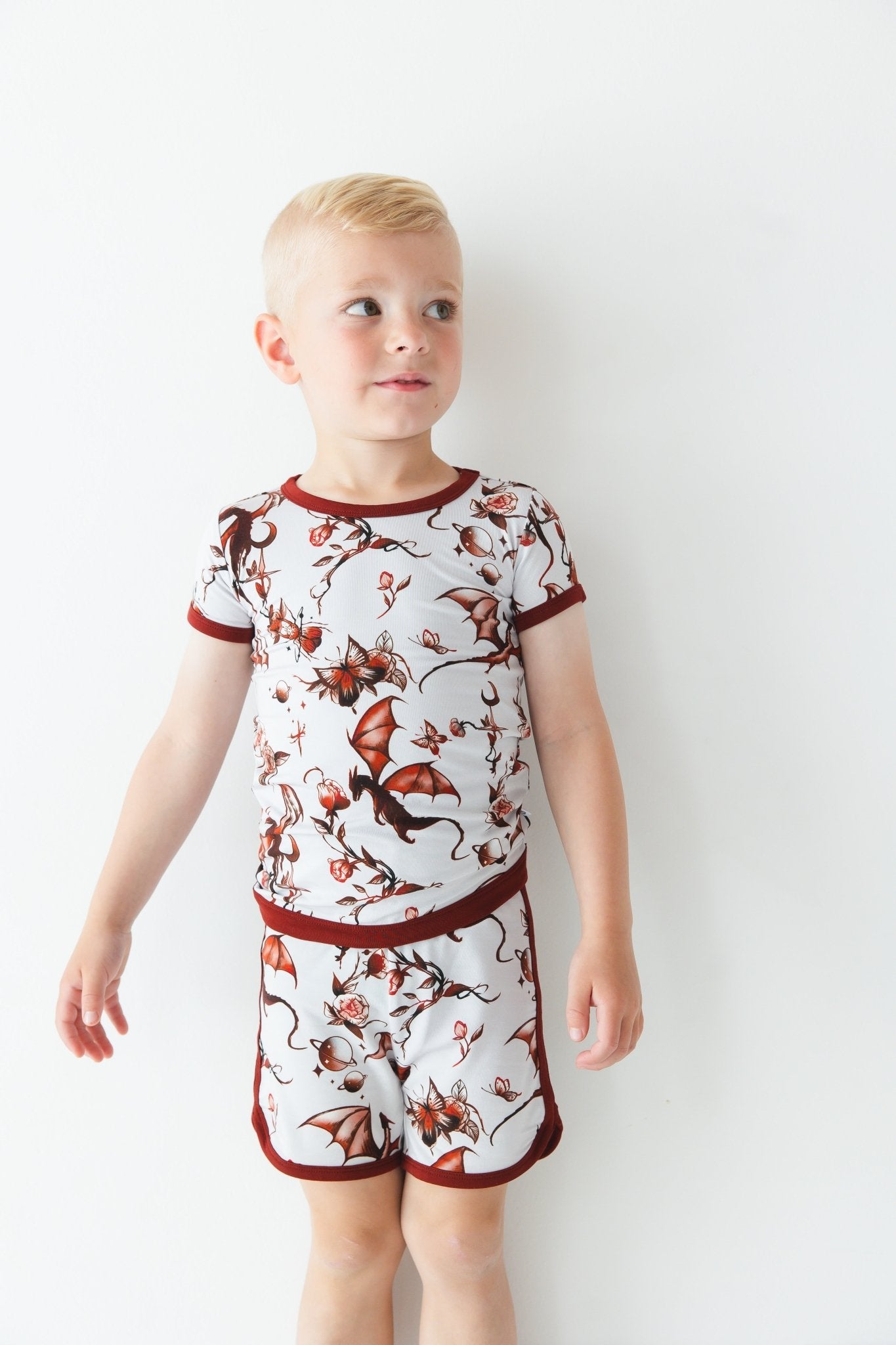 A young child with short blonde hair wears the Jammie Session Dreaming with Dragons Short Pajama Set, featuring a brown dragon and floral print, against a plain white background.