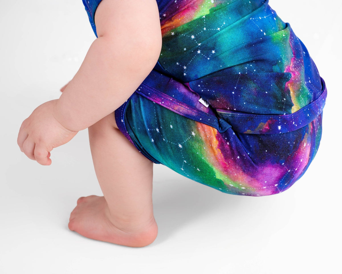 A baby with bare feet crouches in the Jammie Session Luminara Galaxy Short Pajama Set, a colorful romper crafted from buttery soft, hypoallergenic bamboo fabric.