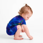 A toddler with light brown hair, wearing the Luminara Galaxy Short Pajama Set by Jammie Session—crafted from buttery soft bamboo in a blue galaxy print—squats on a white floor and reaches toward the ground.