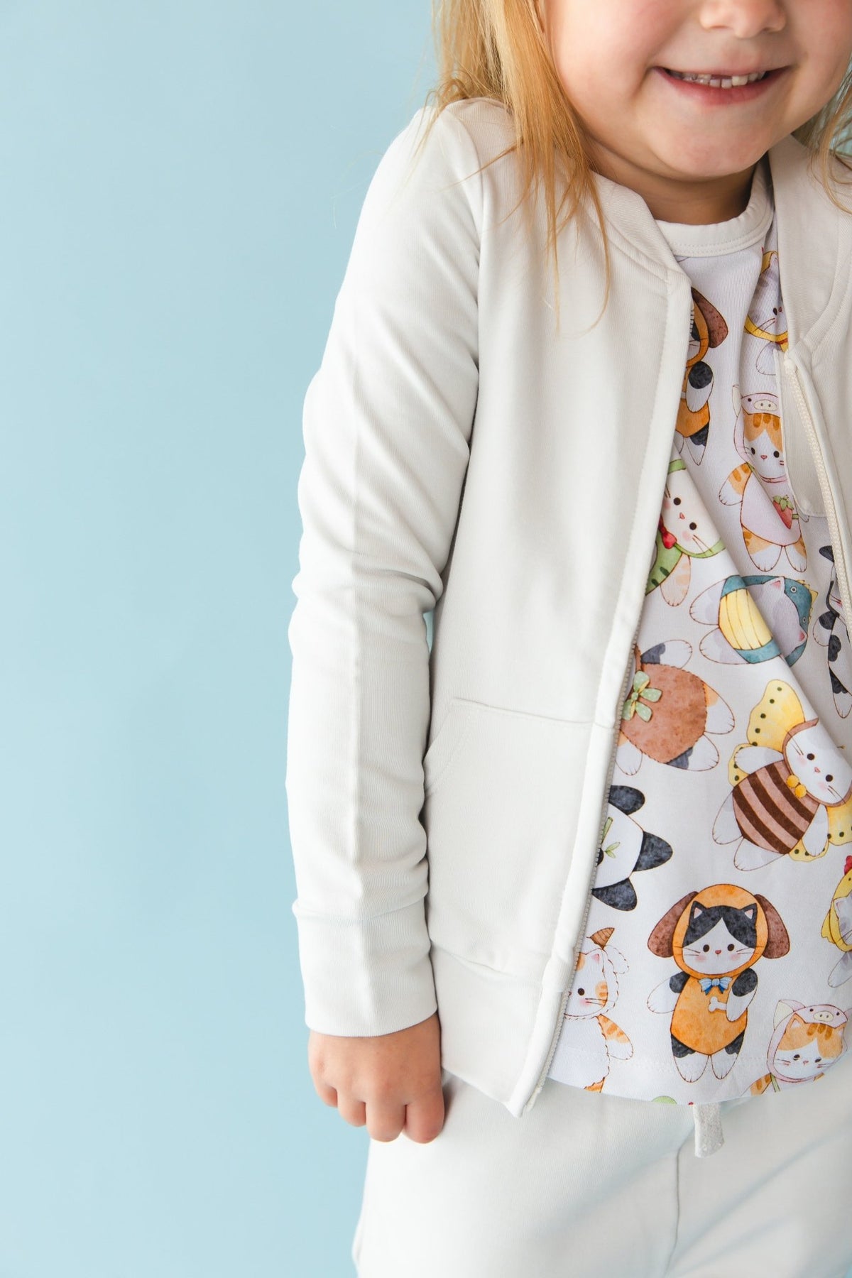 A smiling young child wears the Jammie Session "Meowster of Disguise" Solid Kids' Jacket (runs small, please size up) with matching pants and a cartoon animal print shirt, standing against a light blue background.