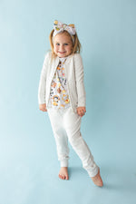 A young girl smiles at the camera, barefoot against a light blue background, wearing the Jammie Session Meowster of Disguise Solid Kids' Jacket (runs small, please size up), a printed shirt, and a matching bow headband.