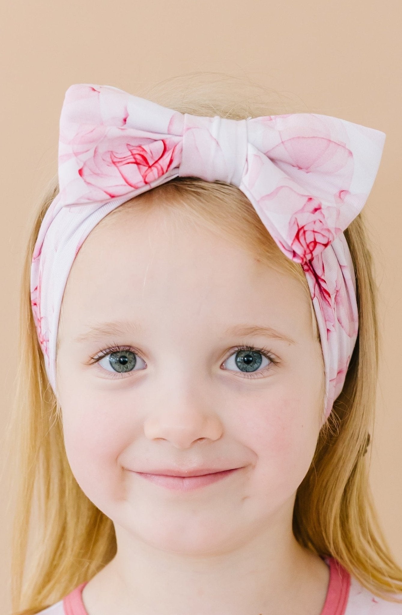A young blonde child wears the Jammie Session Misty Roses Bow pink flower-patterned headband, looking at the camera against a beige background.