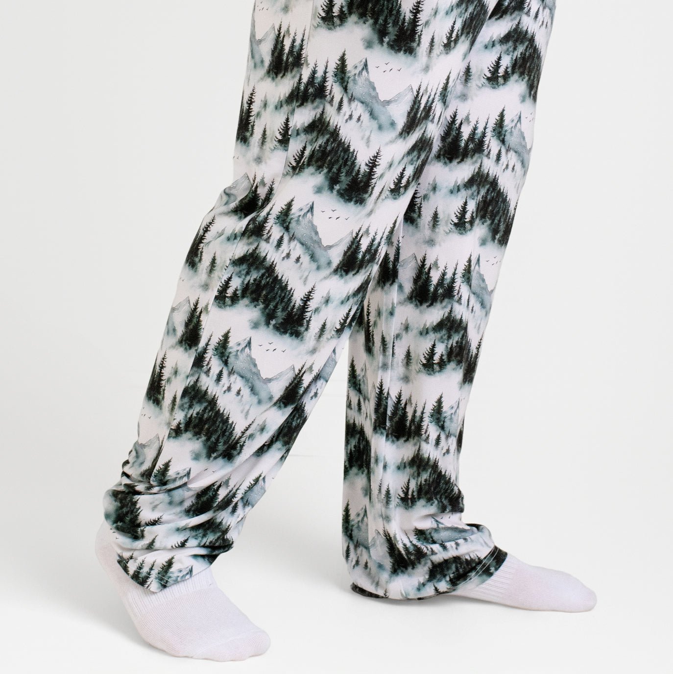 A person wears Jammie Session's Mountain Mist Men's PJ Pants, featuring a soft forest print and made from viscose from bamboo, along with white socks, standing against a plain white background.