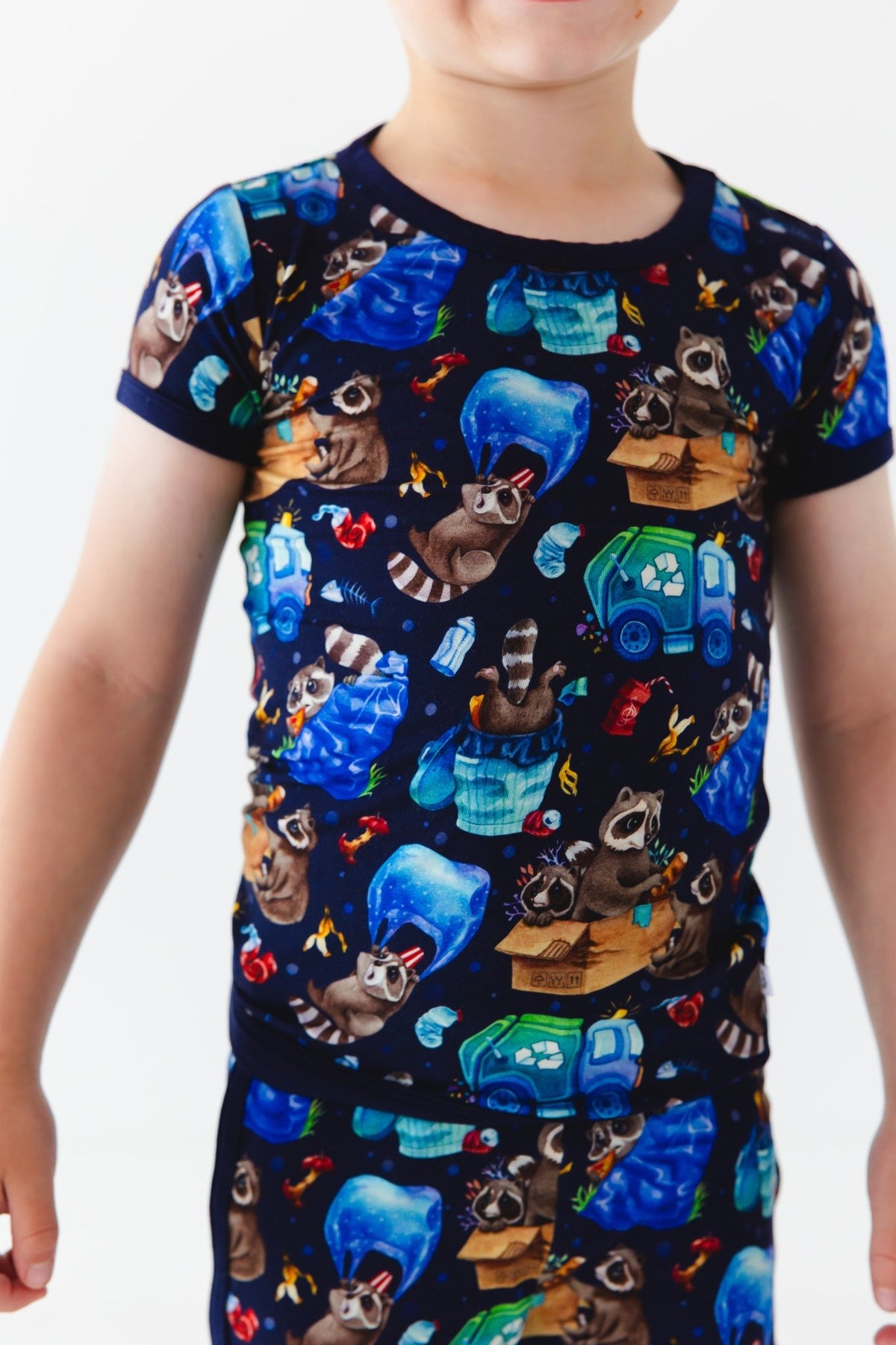 A child wears the Jammie Session Nocturnal Nonsense Short Pajama Set, which features a colorful raccoon and garbage print on navy blue. This hypoallergenic, breathable set keeps kids comfy and stylish all day.