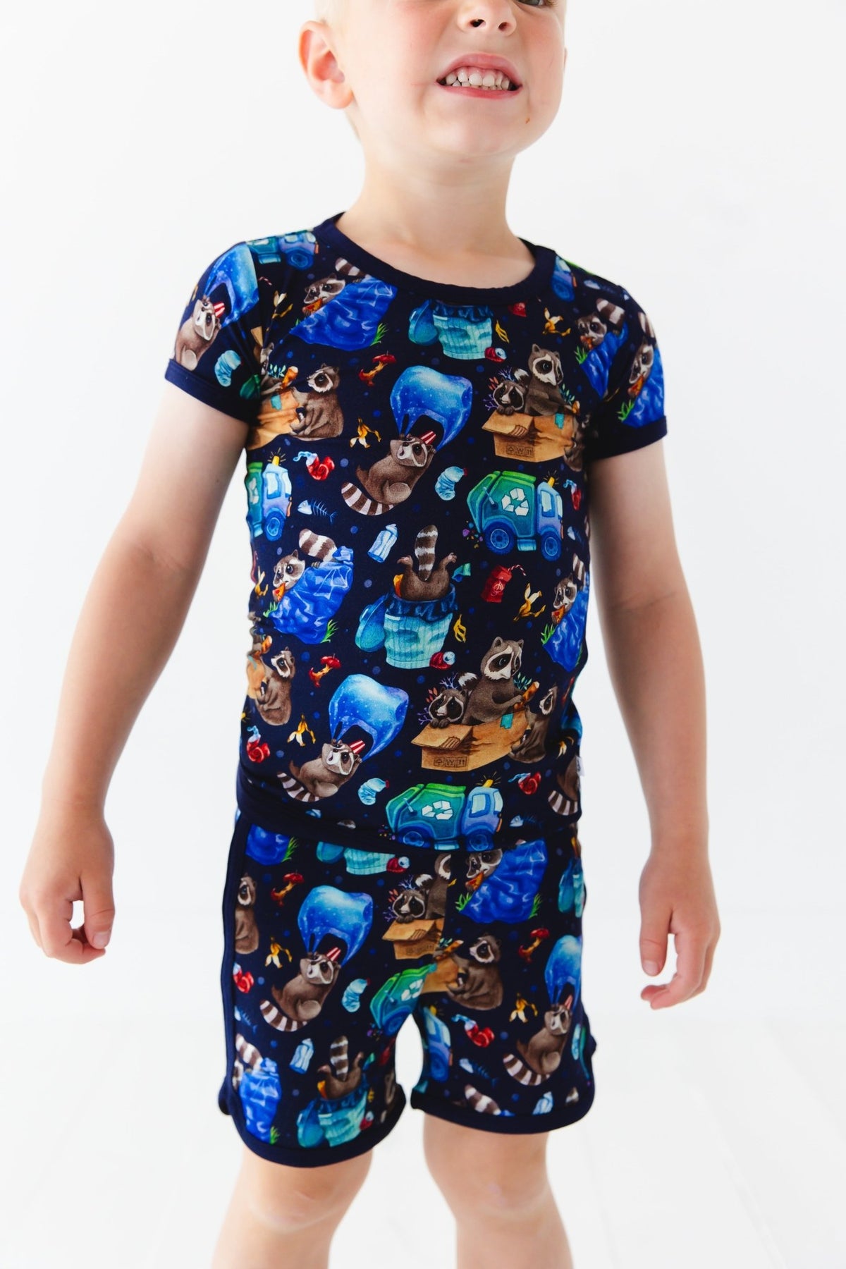 A young child smiles, wearing the Jammie Session Nocturnal Nonsense Short Pajama Set featuring a colorful raccoon and camping-themed print on a dark background.
