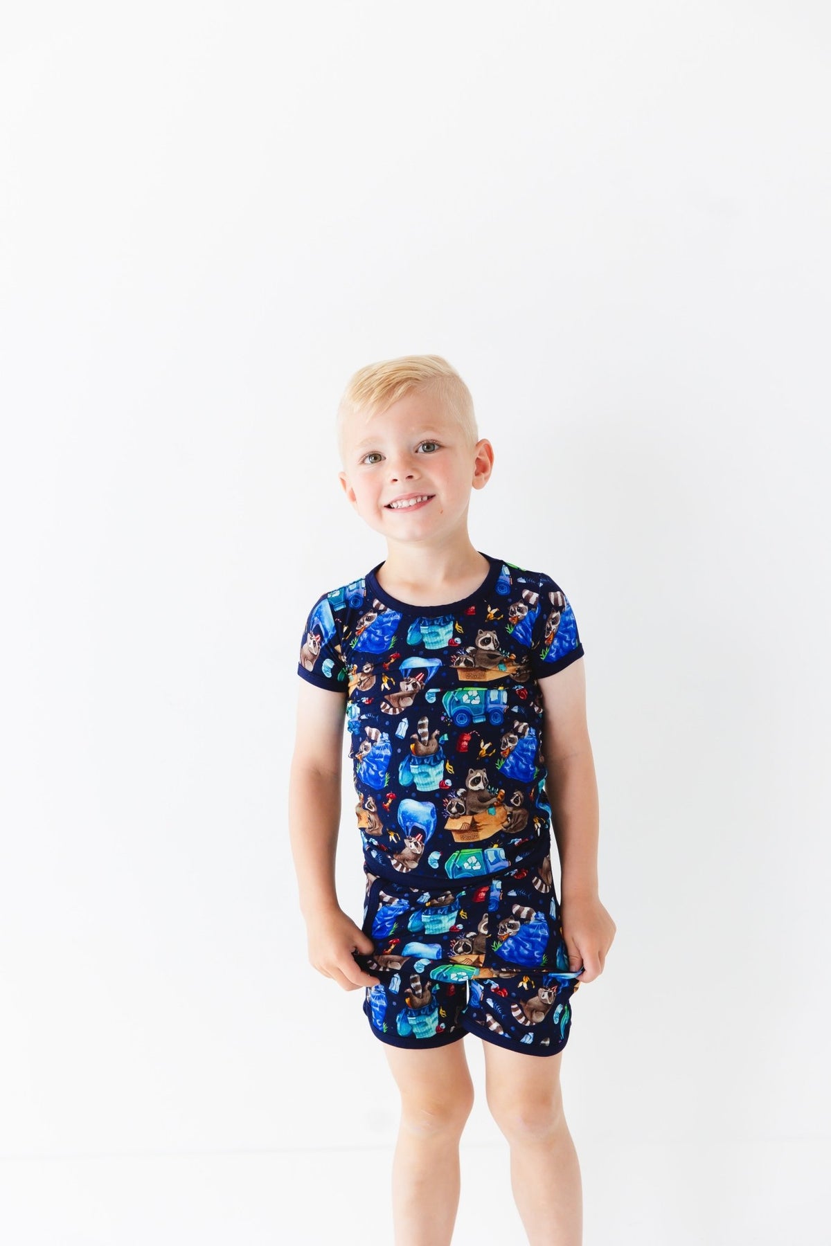 A young boy with short blond hair stands against a plain white background, wearing the Jammie Session Nocturnal Nonsense Short Pajama Set with colorful cartoon animal and space-themed prints.