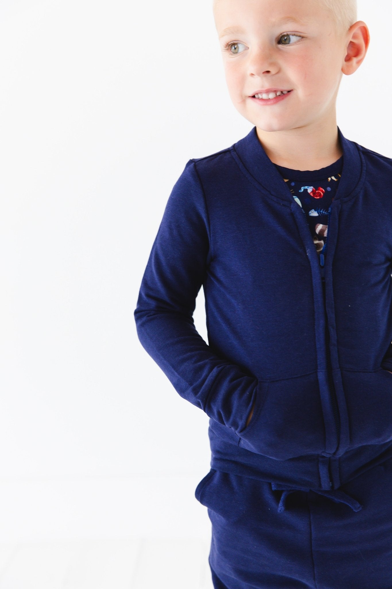 A young child smiles to the side with hands in pockets, wearing a Jammie Session Nocturnal Nonsense Solid Kids' Jacket (runs small, please size up) against a plain white background.