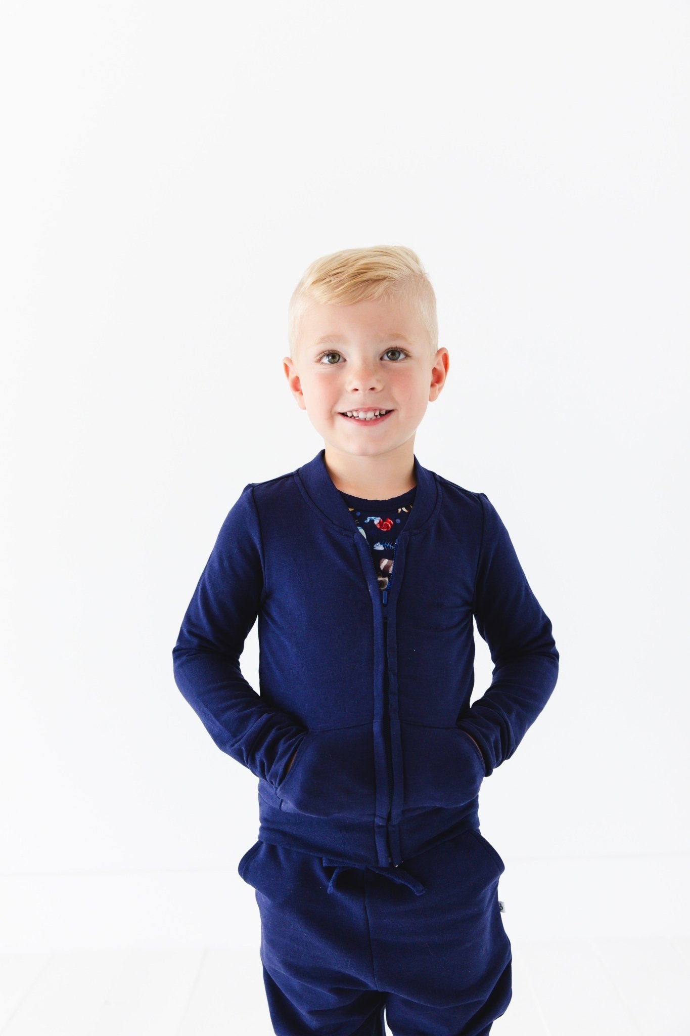 A smiling young child with short blond hair wears the Jammie Session Nocturnal Nonsense Solid Kids' Jacket (runs small, please size up), standing against a plain white background with hands in pockets.