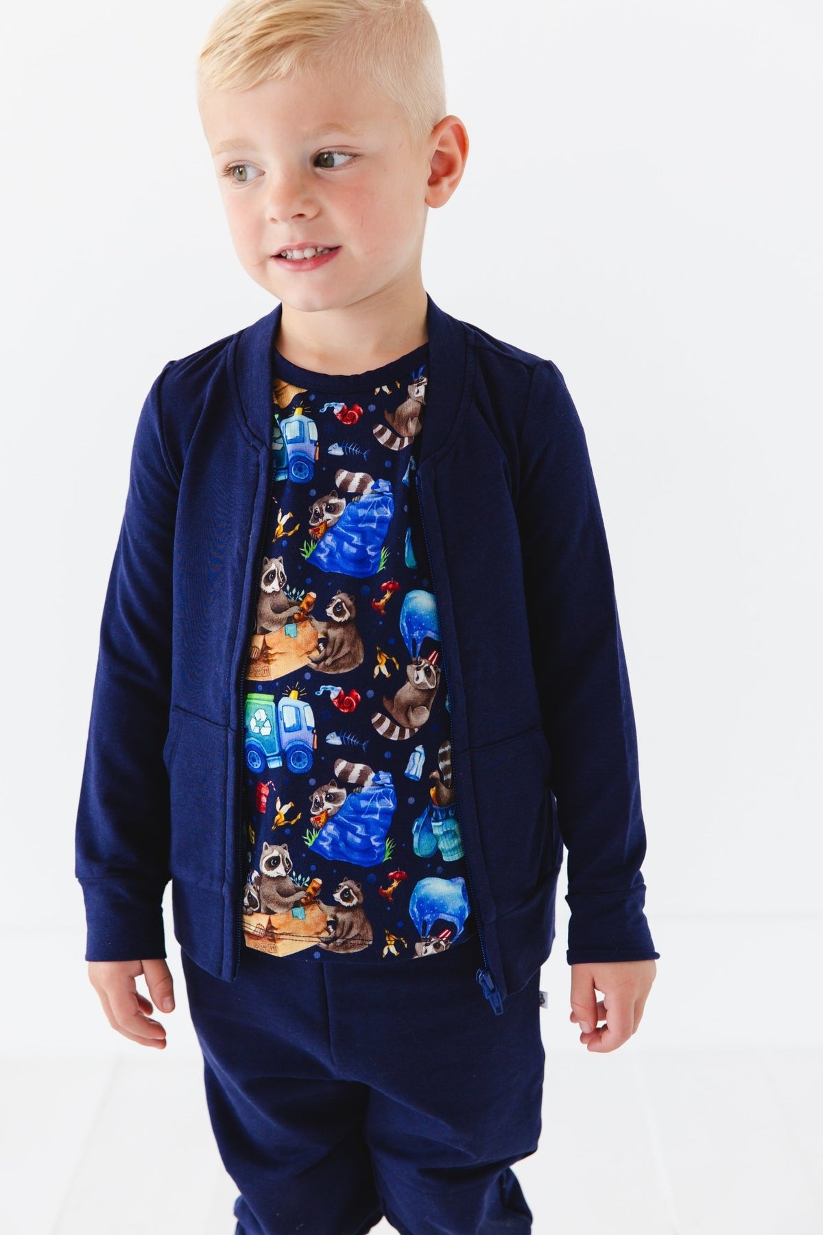 A young child with short blond hair wears the Jammie Session Nocturnal Nonsense Solid Kids' Jacket (runs small, please size up) and pants over a colorful raccoon-print shirt, standing against a white background.