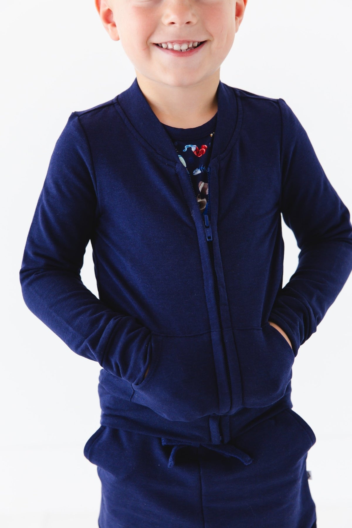 A young child smiles with hands in pockets, wearing the Jammie Session Nocturnal Nonsense Solid Kids' Jacket (navy blue, runs small—please size up), against a white background.