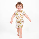 A young child with curly hair smiles barefoot against a plain white background, wearing the Jammie Session Oh Bother, It's Bedtime Short Pajama Set—crafted from buttery soft, hypoallergenic bamboo in a matching nature-print.