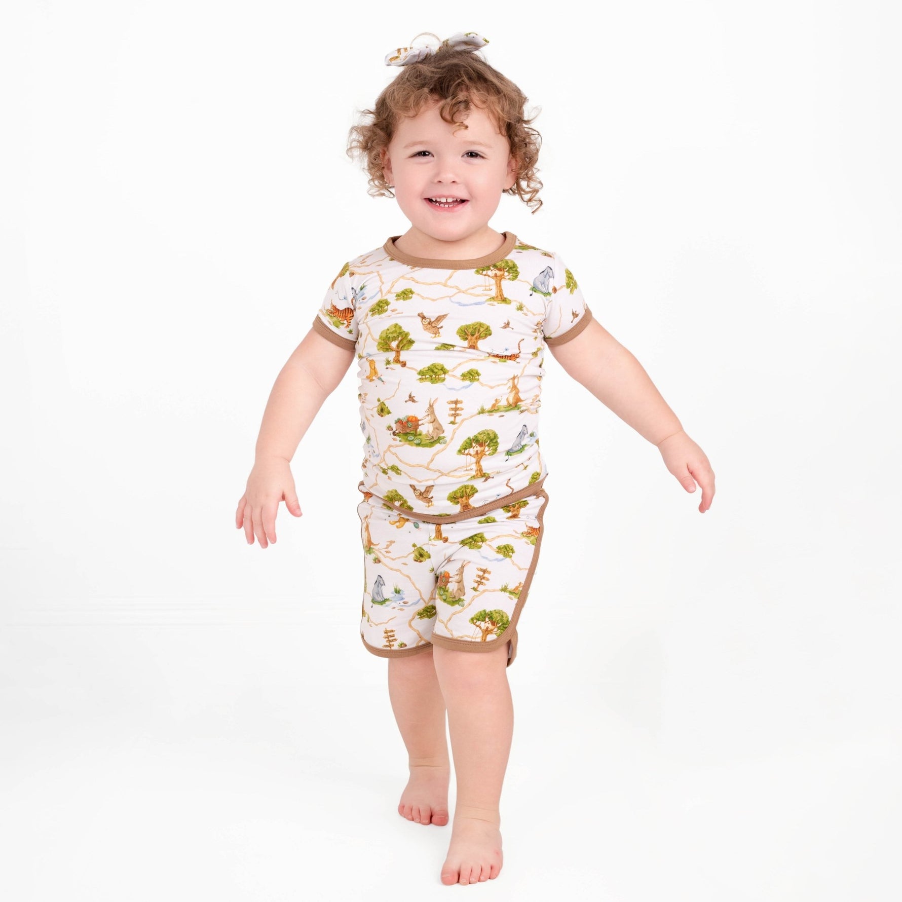 A young child with curly hair smiles barefoot against a plain white background, wearing the Jammie Session Oh Bother, It's Bedtime Short Pajama Set—crafted from buttery soft, hypoallergenic bamboo in a matching nature-print.