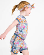A young child wears the Jammie Session Whispering Wildflowers Short Pajama Set—featuring a colorful floral print, matching headband, and made from buttery soft, hypoallergenic bamboo viscose—walking against a plain white background.