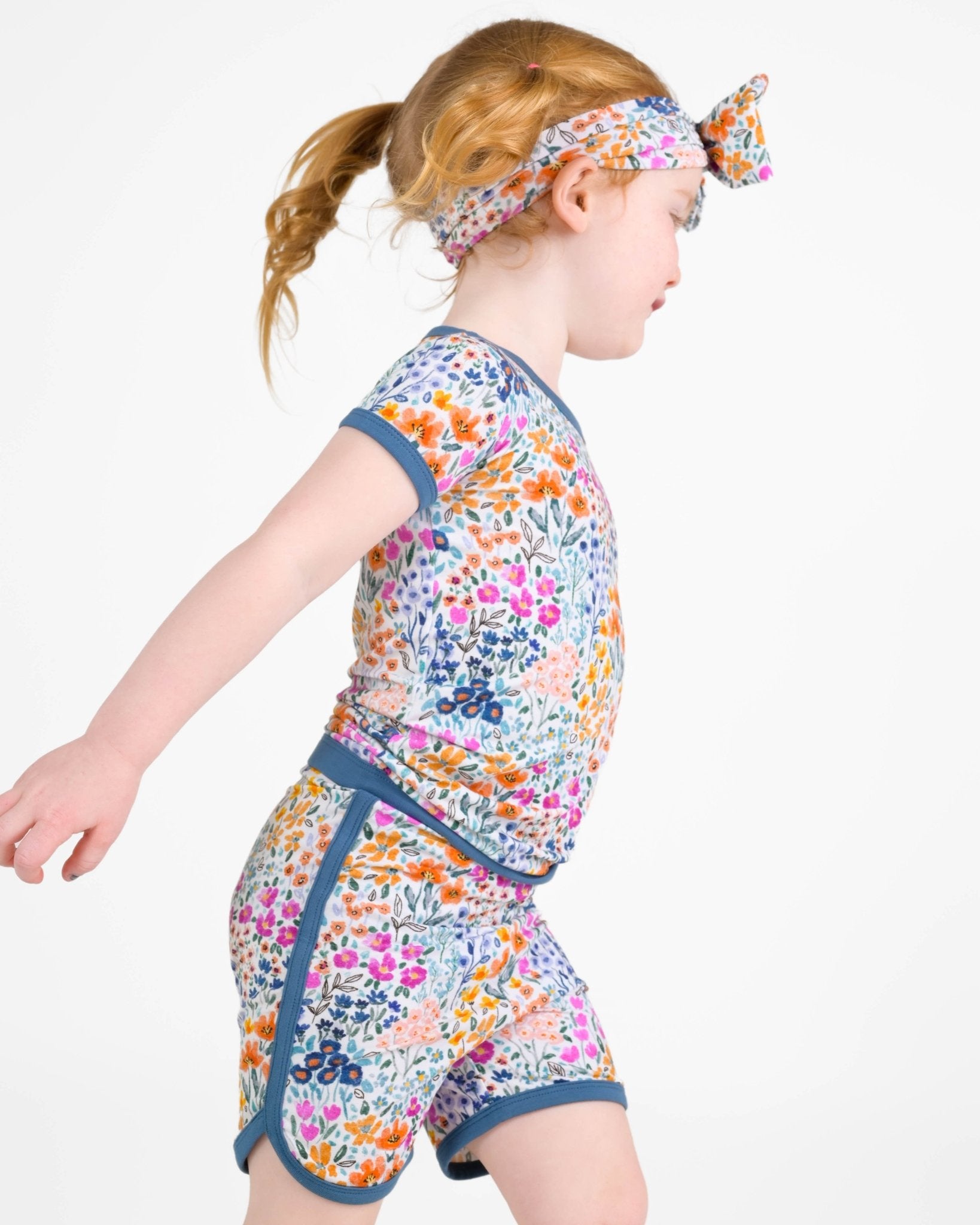 A young child wears the Jammie Session Whispering Wildflowers Short Pajama Set—featuring a colorful floral print, matching headband, and made from buttery soft, hypoallergenic bamboo viscose—walking against a plain white background.