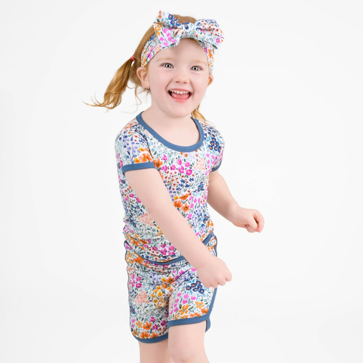 A young child with light hair smiles against a plain white background, wearing the Jammie Session Whispering Wildflowers Short Pajama Set made from buttery soft bamboo viscose, featuring a matching floral shirt, shorts, and headband.