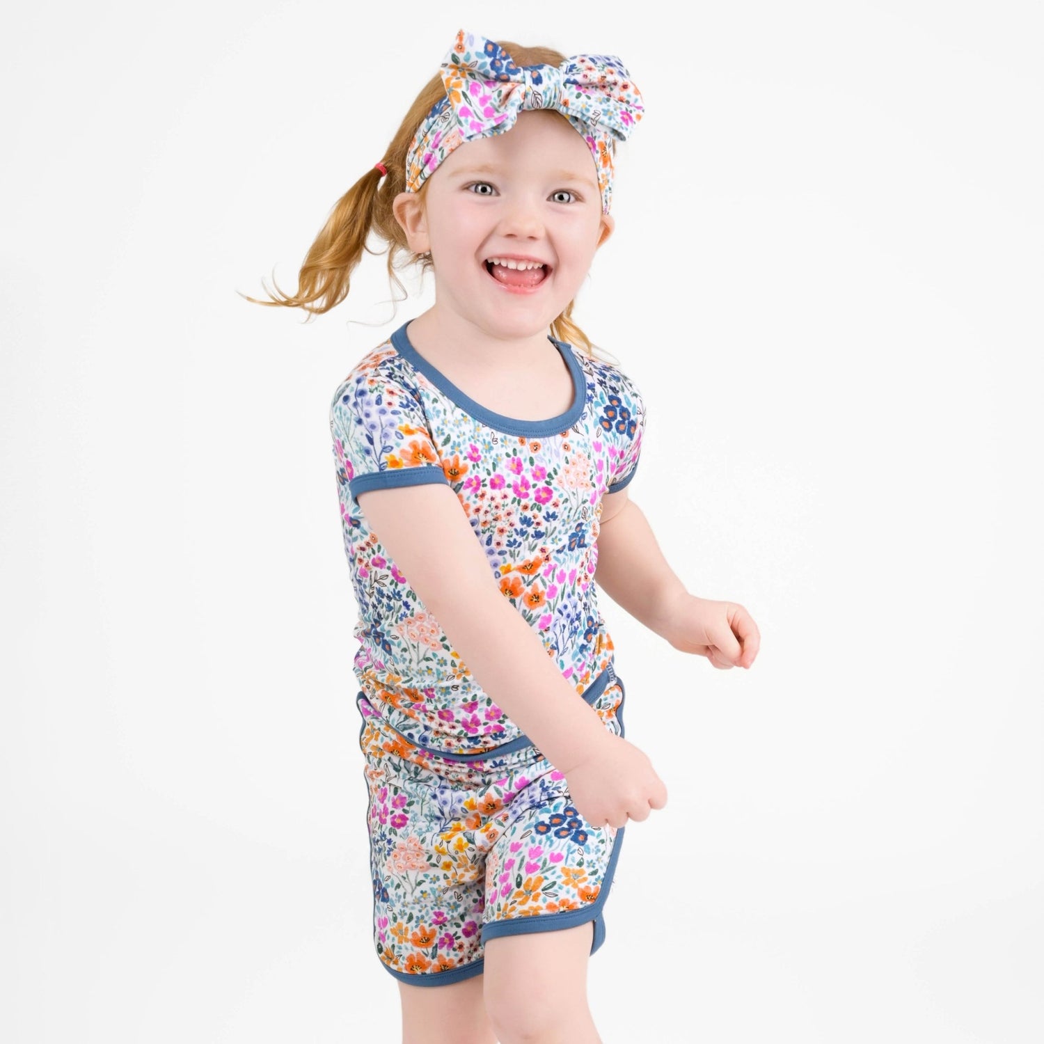 A young child with light hair smiles against a plain white background, wearing the Jammie Session Whispering Wildflowers Short Pajama Set made from buttery soft bamboo viscose, featuring a matching floral shirt, shorts, and headband.