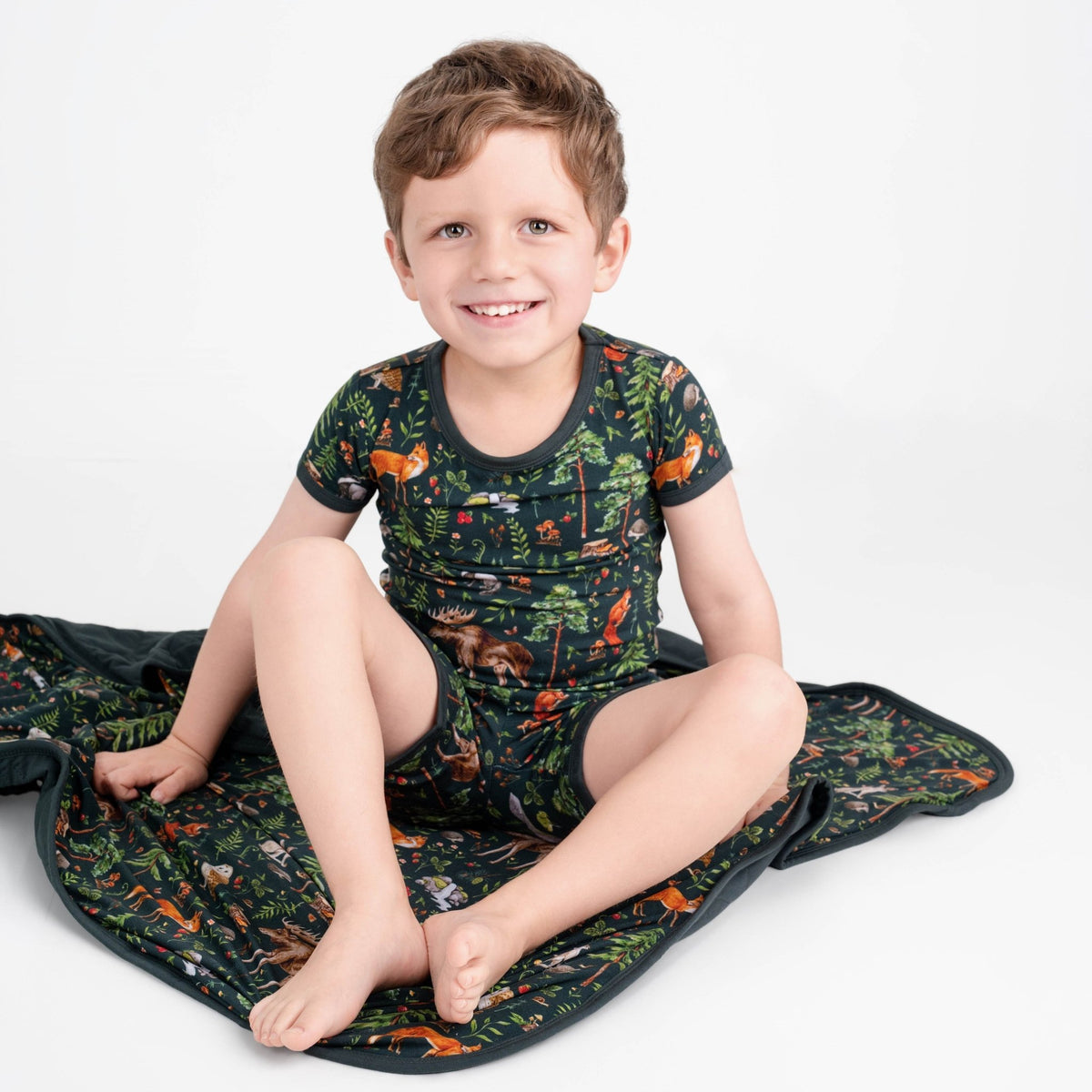 A young boy wears the Jammie Session "Wood You Be My Friend" Short Pajama Set—hypoallergenic bamboo shirt and shorts with a colorful animal and forest print—while sitting on a matching blanket against a plain white background.