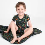 A young boy wears the Jammie Session "Wood You Be My Friend" Short Pajama Set—hypoallergenic bamboo shirt and shorts with a colorful animal and forest print—while sitting on a matching blanket against a plain white background.