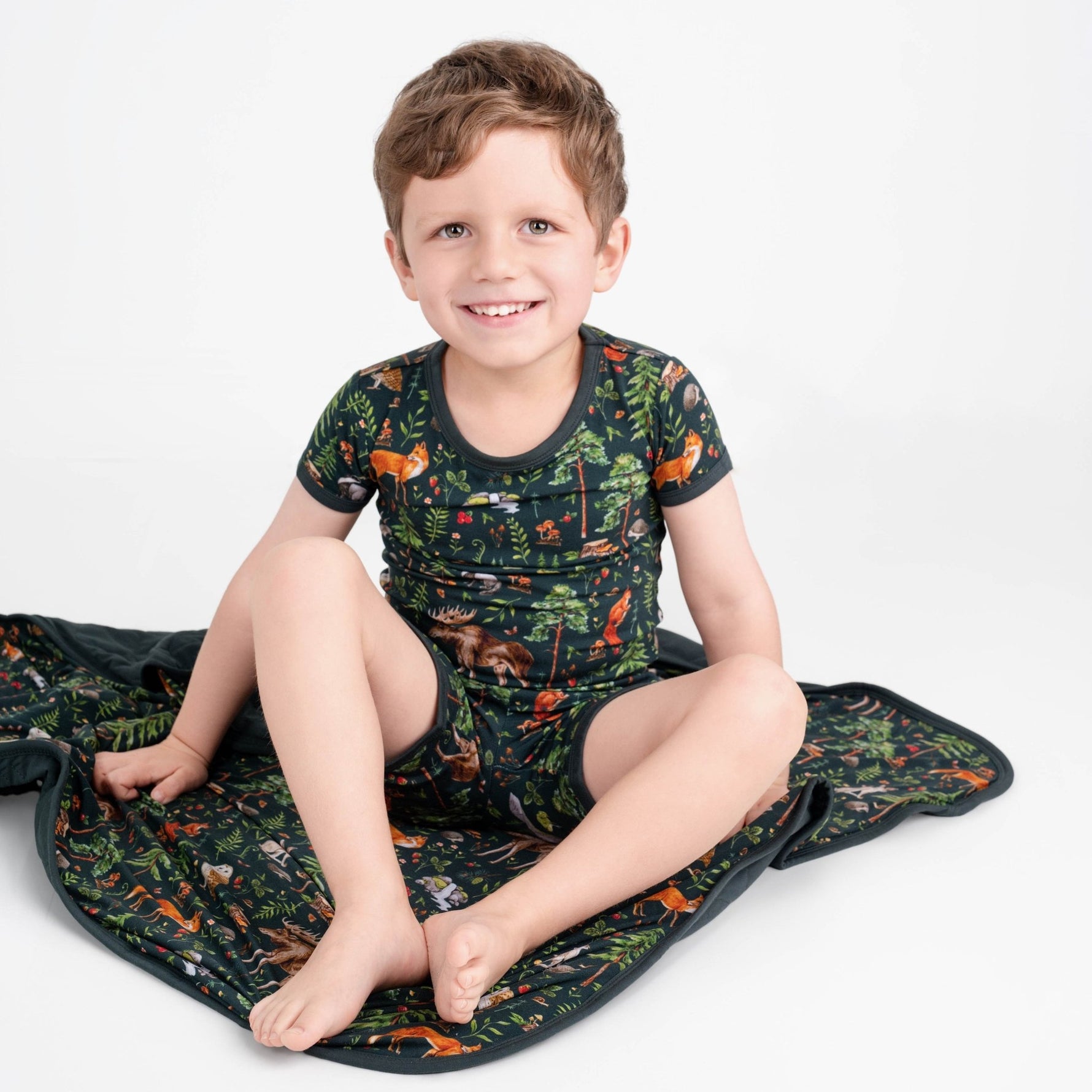 A young boy wears the Jammie Session "Wood You Be My Friend" Short Pajama Set—hypoallergenic bamboo shirt and shorts with a colorful animal and forest print—while sitting on a matching blanket against a plain white background.