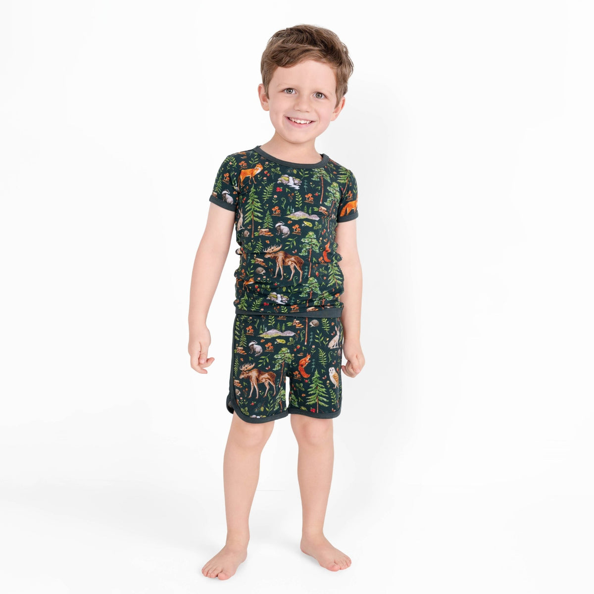 A young boy smiles barefoot in Jammie Session’s "Wood You Be My Friend" Short Pajama Set, featuring a dark short-sleeve shirt and shorts with colorful forest animals on white, made from hypoallergenic, moisture-wicking bamboo for comfort.