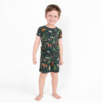 A young boy smiles barefoot in Jammie Session’s "Wood You Be My Friend" Short Pajama Set, featuring a dark short-sleeve shirt and shorts with colorful forest animals on white, made from hypoallergenic, moisture-wicking bamboo for comfort.