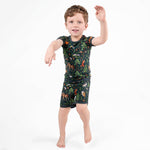 A young boy poses on one foot with arms raised, wearing the Jammie Session Wood You Be My Friend Short Pajama Set—moisture-wicking bamboo clothing in a dark jungle print, perfect for active kids—against a plain white background.