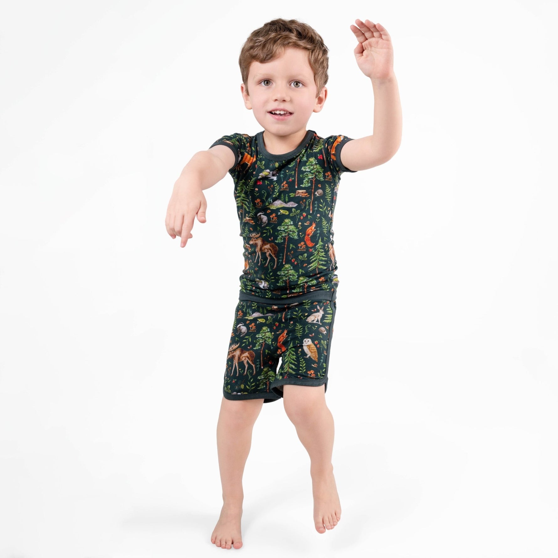 A young boy poses on one foot with arms raised, wearing the Jammie Session Wood You Be My Friend Short Pajama Set—moisture-wicking bamboo clothing in a dark jungle print, perfect for active kids—against a plain white background.