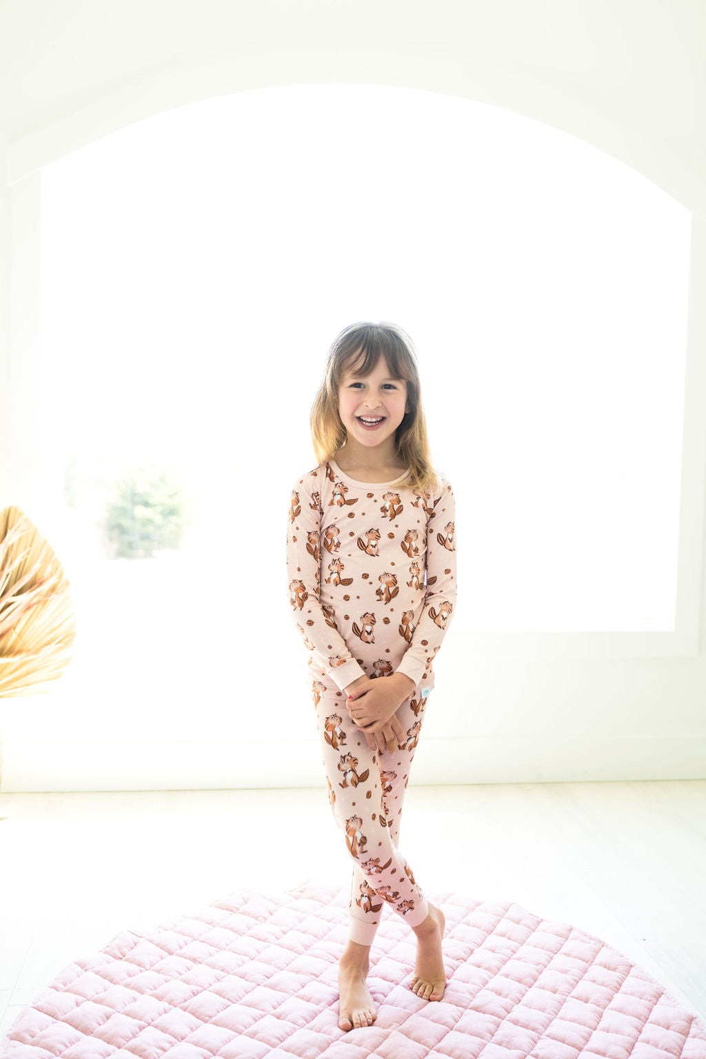 A young girl stands barefoot on a pink quilt, smiling and wearing Alvin Long Sleeve PJ's BDLJ by Big Dreams Little Jammies, featuring a brown animal print, in a bright white room.